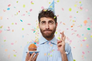 Un chico joven con barba con un pastel con vela en una mano y cruzando las manos de la otra mira hacia el cielo en una supuesta fiesta de cumpleaños
