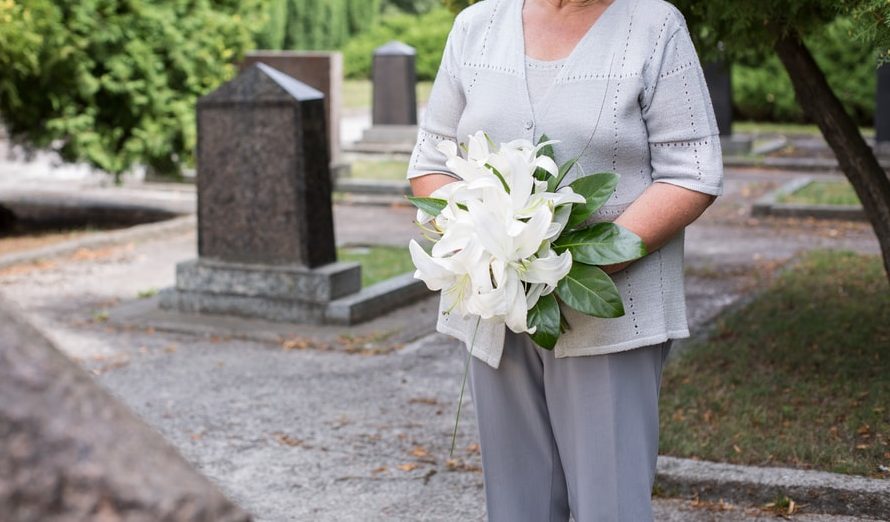 Una mujer de edad avanzada sujeta un ramo de lirios blancos frente a una tumba